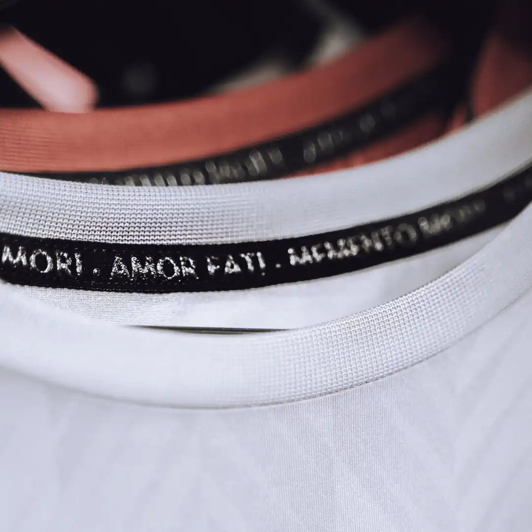 Clothing tag with text ’MORT. AMOR FATI’ printed on black fabric against white material.