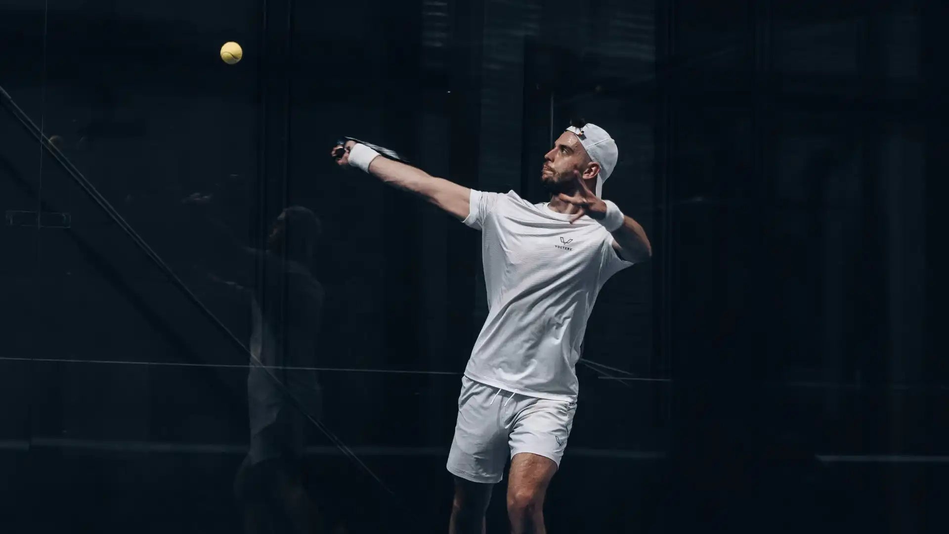 Tennis player serving a ball while wearing light-colored athletic attire.